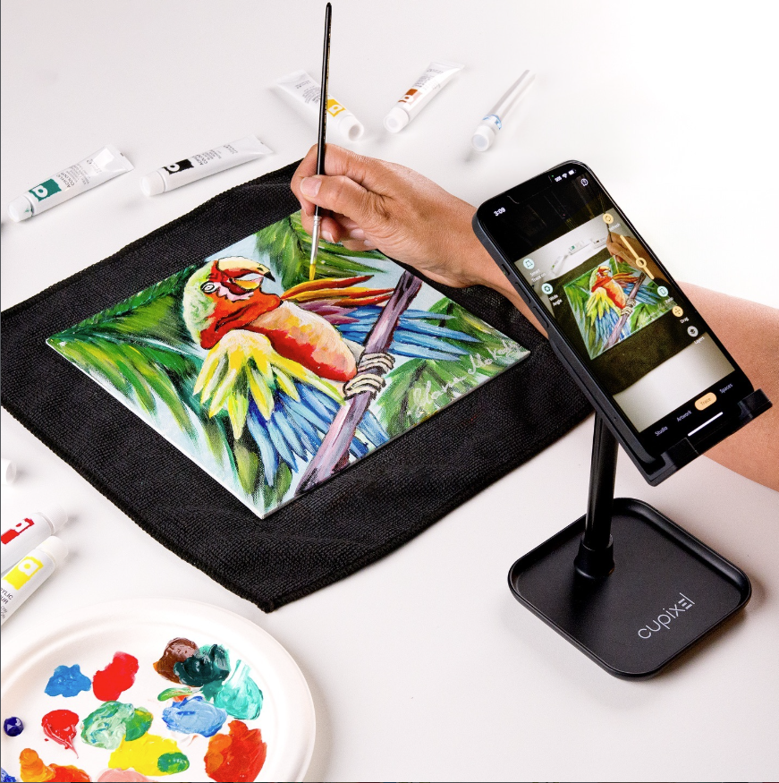 Cupixel phone stand showing a painting reference while a user paints a colorful parrot on a real canvas